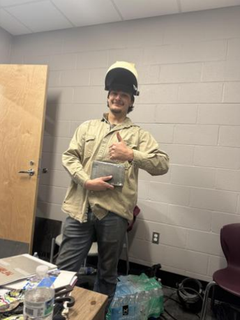    A student stands in a classroom wearing a flipped‑up welding helmet and a beige protective welding jacket. They hold a rectangular metal piece in one hand and give a thumbs‑up with the other. Behind them is a light gray cinder block wall, an open wooden door, and a table with a laptop, water bottle, and assorted supplies. Additional bottles and containers sit on the floor along the wall. The scene captures a moment of hands‑on welding or shop‑class work.
